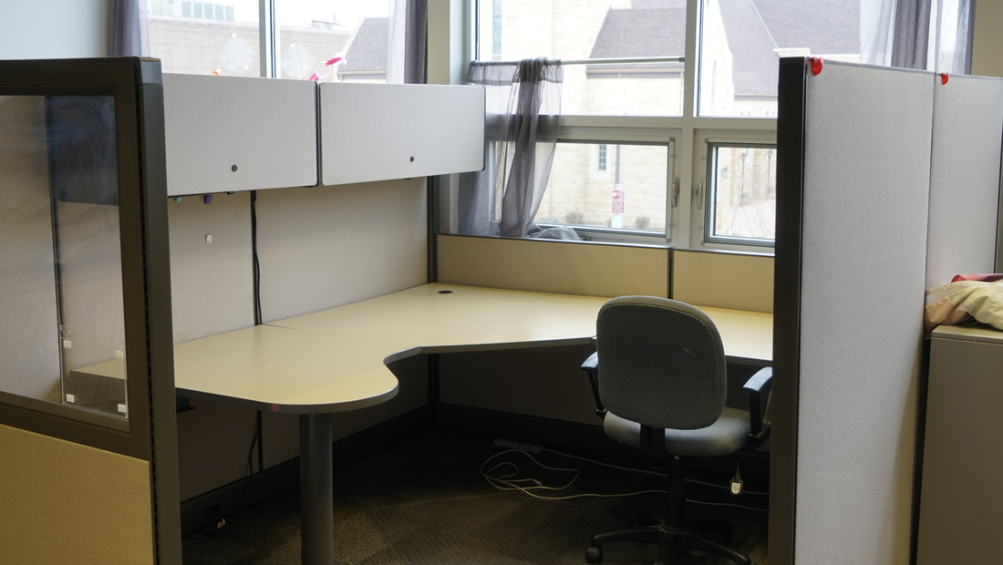 A desk in a corner of windows with a chair pushed in and 2 cabinets above.