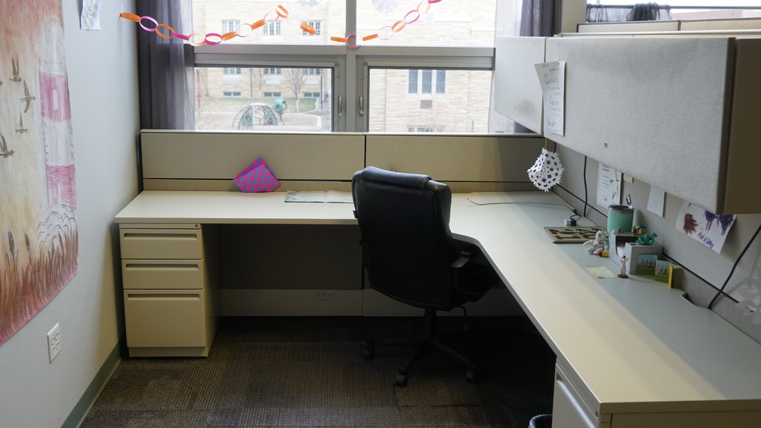A desk space next to a window has an office chair pushed in. There are 2 cabinets above and below are 3 drawers on both sides of the desk.